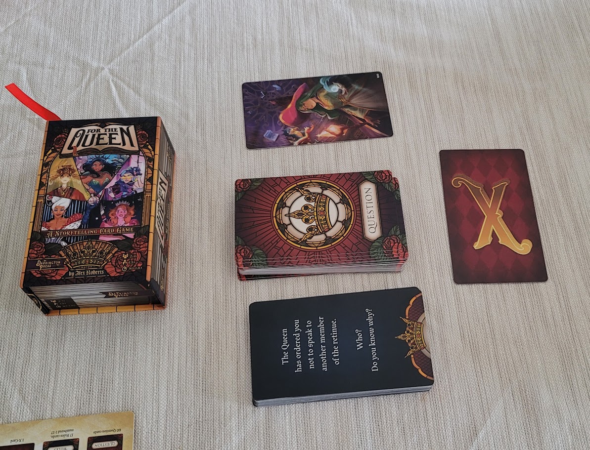 The box for the game For the Queen, across from the X-Card opposite the box. In the middle are a stack of question cards, with a discard pile on one side and the image of The Witch Queen on the other side.
