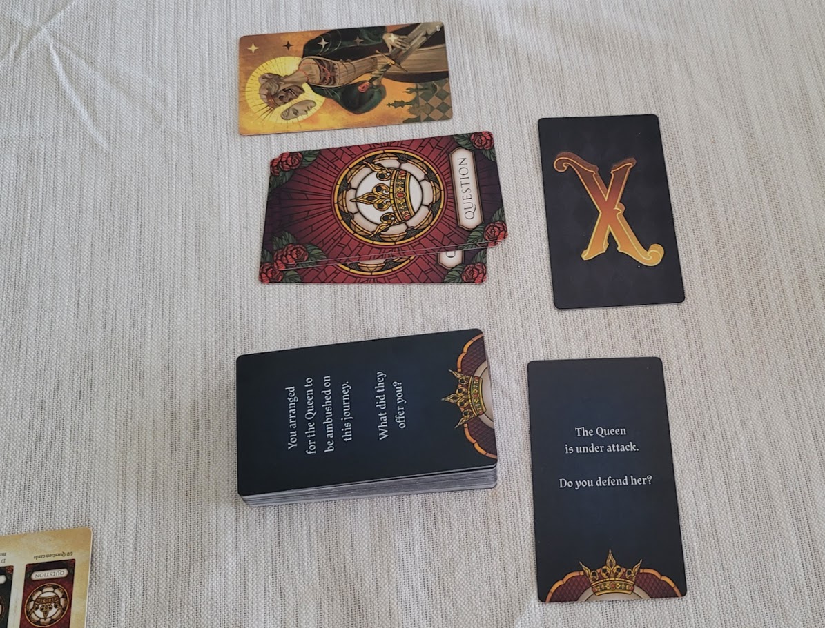 Question cards in the middle of the table, with a discard pile on one side, and the card with the image of the Undead Queen on the other side. There is an X-Card sitting to one side, above another card that reads "The Queen is Under Attack. Do You Defend Her?"